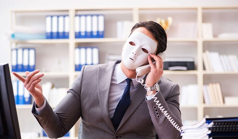 Mann im Anzug mit weißer Maske telefoniert im Büro, zeigt mit der Hand; viele Regale im Hintergrund.