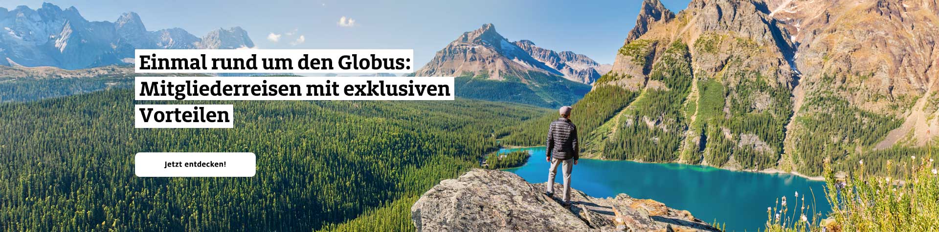 Ein Wanderer steht auf einem Felsen, blickt auf türkisfarbenen See und Berge unter blauem Himmel.