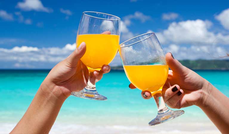 Zwei Gläser mit Orangensaft vor blauem Meer, beide Hände stoßen an, sonniger Strandhintergrund.